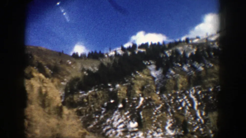 Late clip frame from the 1961 Aspen, Colorado drive, showing a tree-lined slope with patchy snow beneath a bright blue sky.