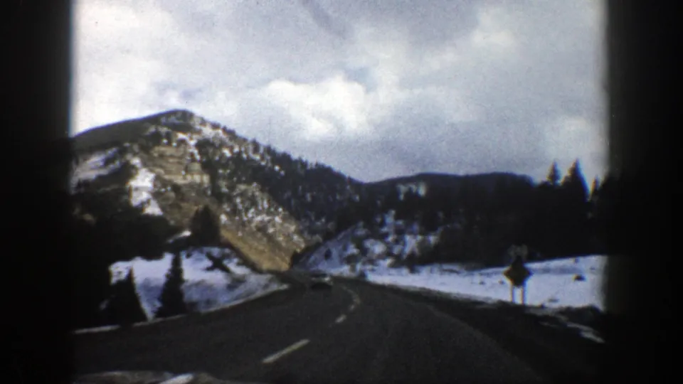 1961 Aspen-area road view from a moving vehicle, with a curve ahead, snowy shoulders, evergreens, and cloudy mountains.
