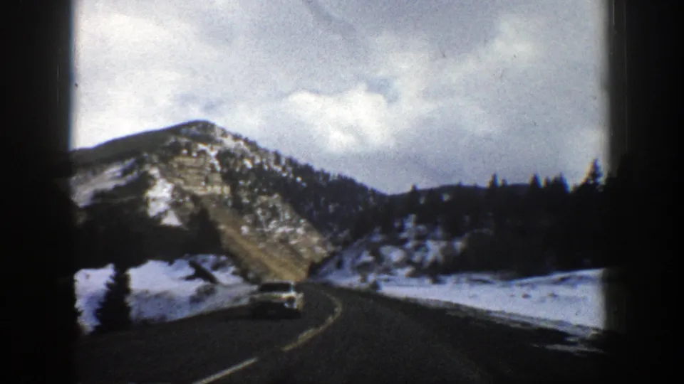 1961 Aspen-area 8mm frame showing a curving road, an oncoming vehicle, snow patches, trees, and a rocky mountain slope.