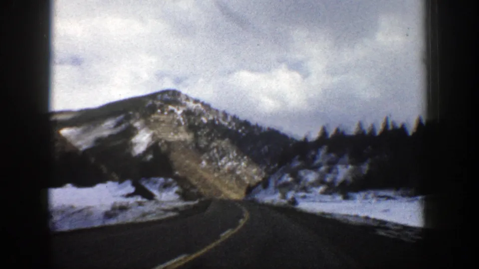 1961 Aspen-area 8mm road scene with snowy shoulders, evergreen silhouettes, rocky slopes, and an overcast mountain sky.