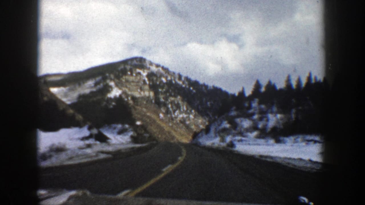 Two-lane mountain road leading toward snowy hills and evergreen trees under a cloudy sky.