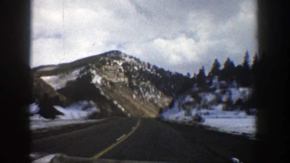 In-car 8mm view of a winding road through snowy Aspen-area mountain terrain, with rocky slopes and evergreens visible.