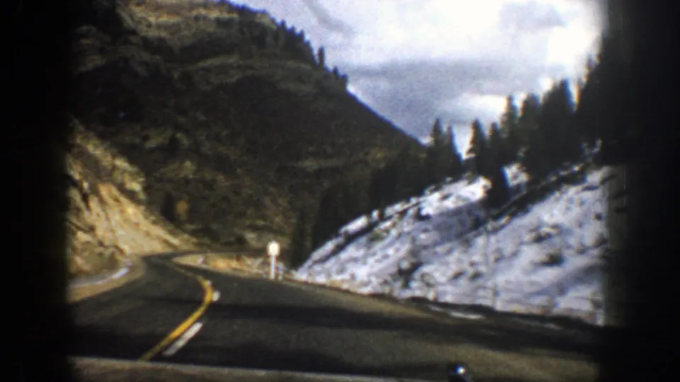 1961 Aspen, Colorado home-movie frame from inside a car, looking down a curving mountain road bordered by snow, trees, and rocky slopes.