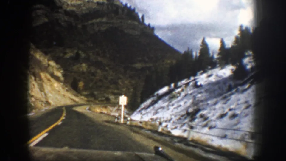 Passenger-view 8mm frame of a narrow Aspen mountain road in 1961, with a left curve, snow patches, evergreen trees, and a roadside sign.