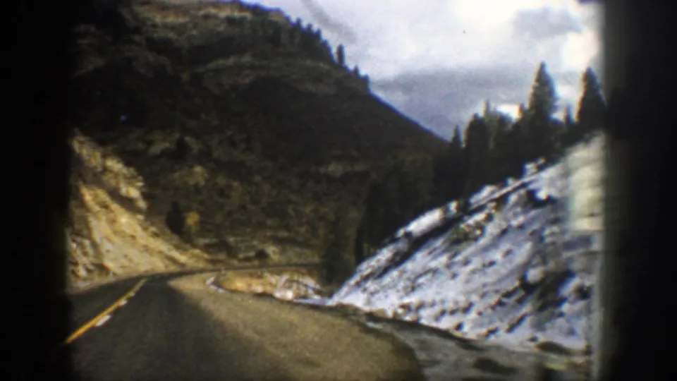 Aspen, Colorado road-travel frame showing a winding asphalt lane, snow along the shoulder, rocky terrain, and conifers under muted light.