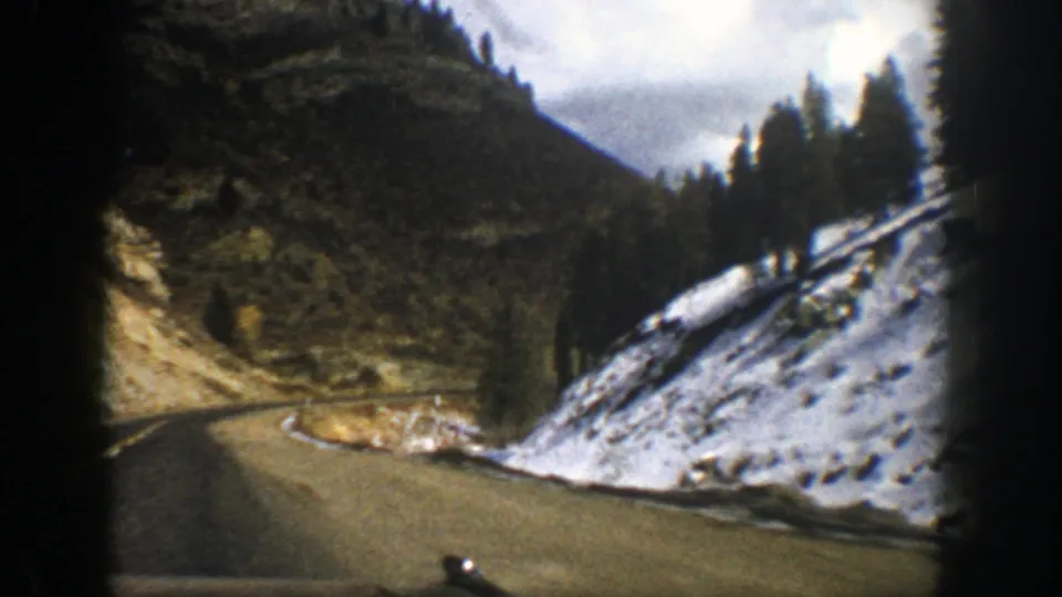 Late-frame view from the moving car on a 1961 Aspen mountain road, with snow on the shoulder, dark trees, and a bend disappearing ahead.
