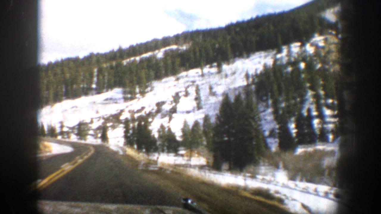 1961 Aspen-area home-movie view of a paved mountain road curving past snow-covered slopes and evergreen trees.