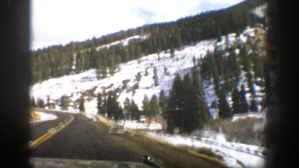 1961 Aspen-area home-movie view of a paved mountain road curving past snow-covered slopes and evergreen trees.
