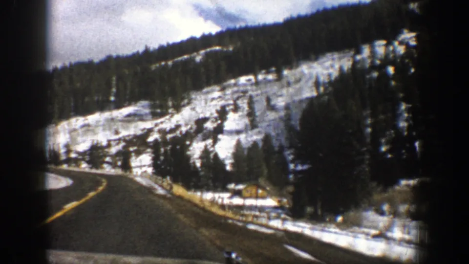 1961 Aspen-area road view with snow-dusted mountainside, evergreens, fencing, and a small roadside structure.