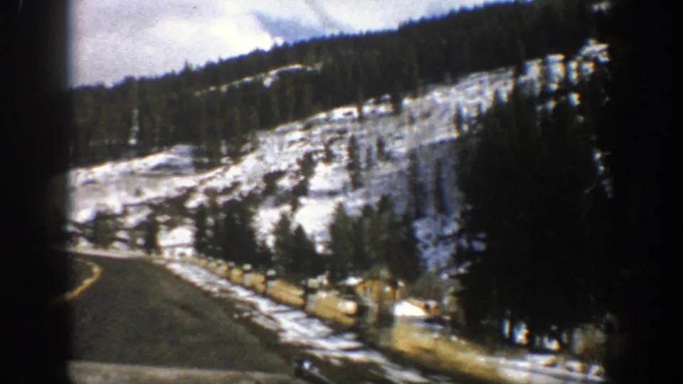 1961 Aspen-area mountain-road frame showing a vehicle view past snow, trees, fencing, and a distant roadside building.