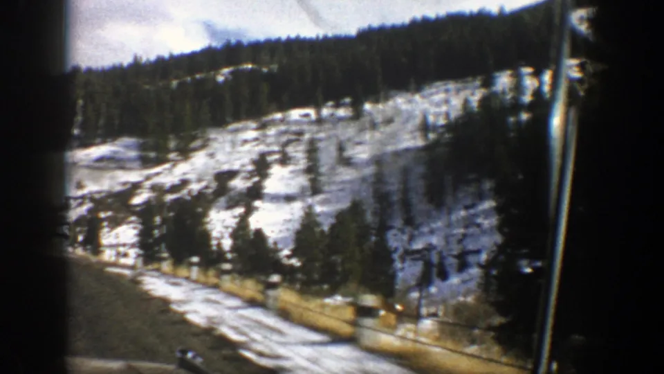 1961 Aspen-area home-movie frame looking from a vehicle toward a snowy slope, road edge, trees, and roadside fencing.