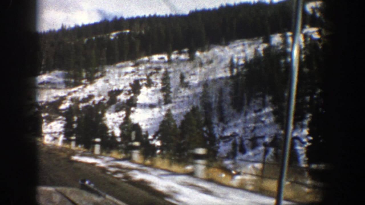 Snow-dusted mountain slope above a paved roadside seen from a moving car.