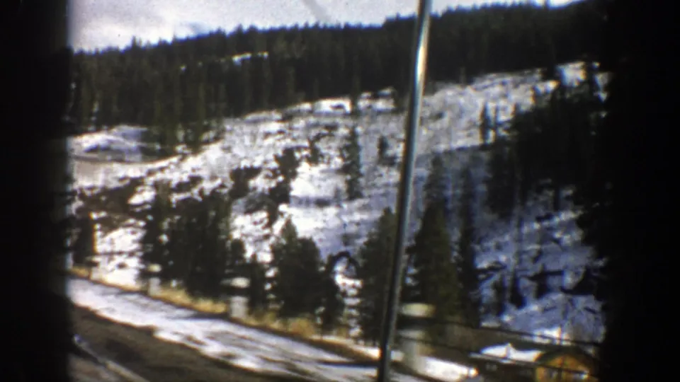 Moving-car view of a snow-patched Aspen mountain slope with evergreen trees and a roadside pole on 8mm film.