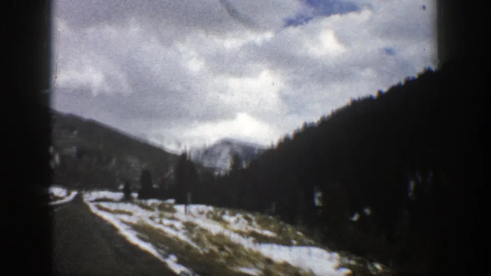 1961 Aspen road scene showing a winding paved road, snow patches, dark evergreen slope, and distant cloudy mountains.