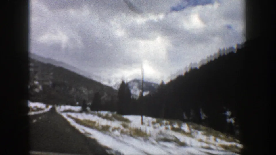 Aspen, Colorado mountain road view with patchy roadside snow, a utility pole, forested hills, and cloudy sky on 8mm film.