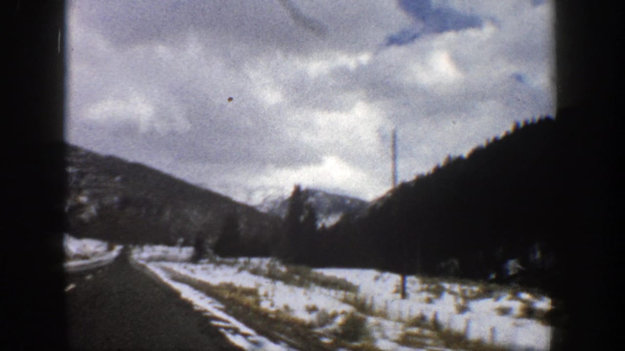 Snowy two-lane road curves through dark mountain slopes under a cloudy sky.