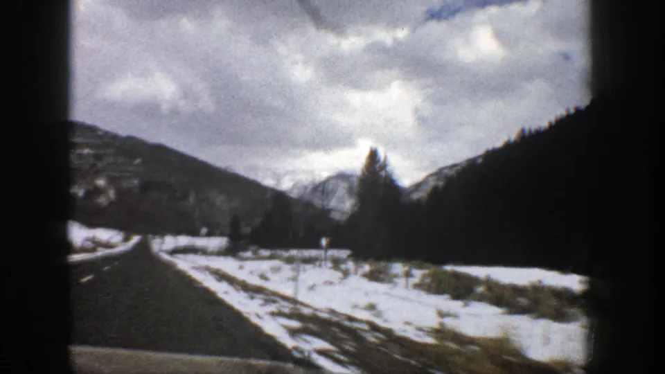 1961 Aspen-area winter road view with snow along both sides, low mountain slopes, bare trees, and a cloudy sky.
