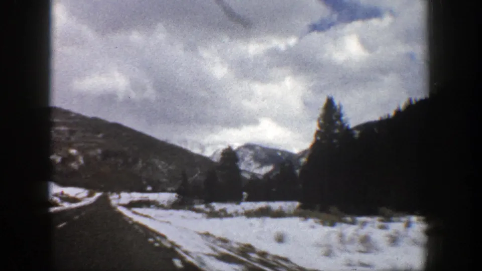 1961 Aspen-area winter drive with a road edge, snowy ground, dark evergreen hillside, bare trees, and distant mountains.