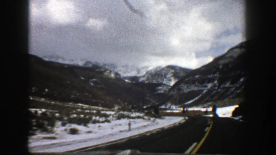 1961 Aspen-area winter road scene with a curving paved route, snowbanks, steep slopes, distant snowy peaks, and cloudy light.