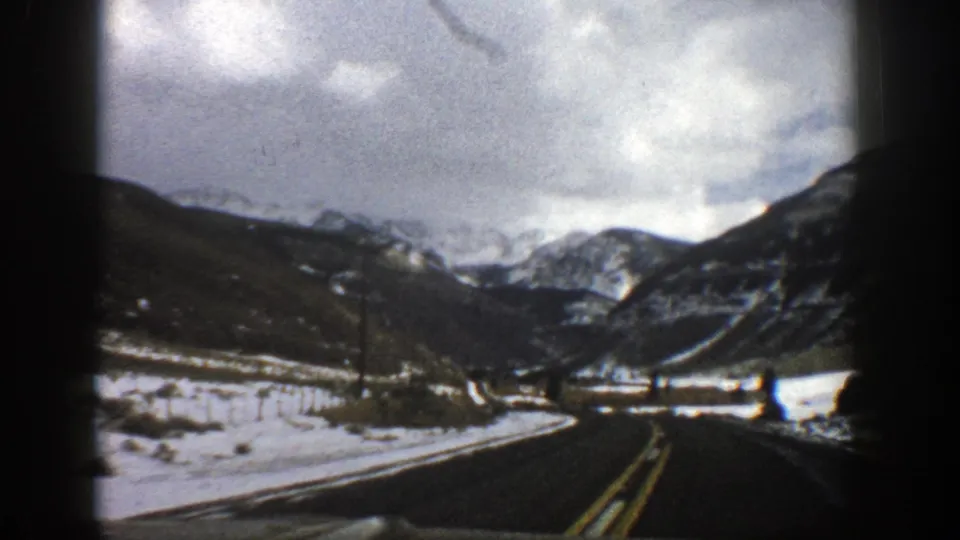 1961 Aspen, Colorado road view from a moving vehicle, with snow patches, utility poles, and mountain peaks ahead.