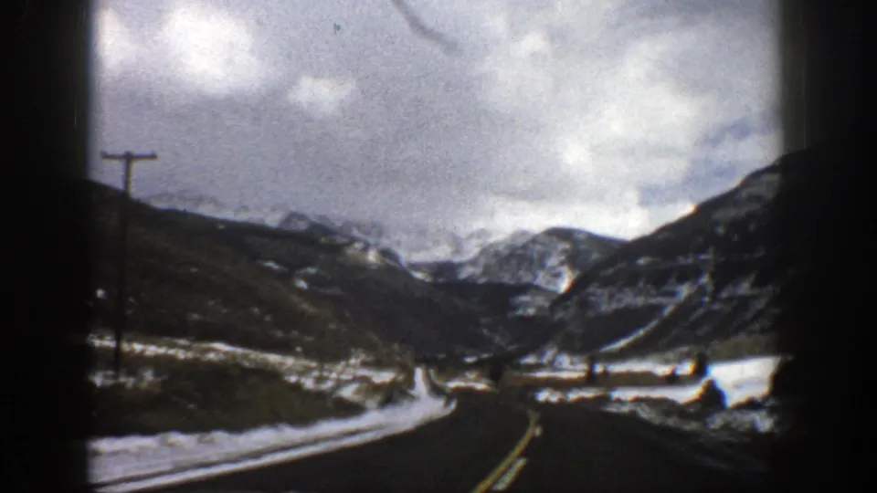 1961 Aspen, Colorado home-movie frame showing a winter road, roadside pole, and snow-streaked mountains from inside a moving car.