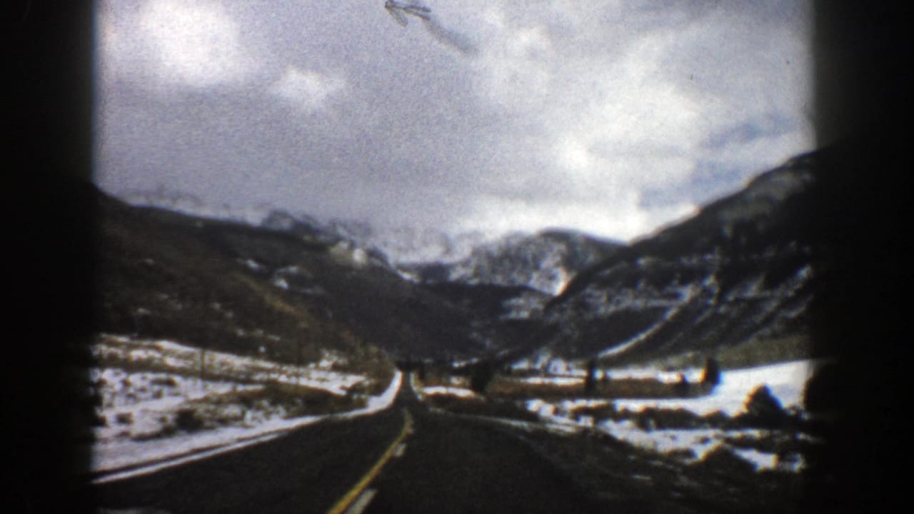 Two-lane road leading toward snowy mountains under cloudy sky.