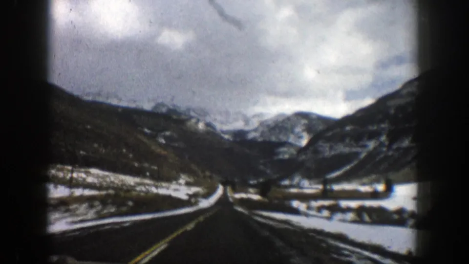 Vintage 8mm road view in Aspen, Colorado, with snow along the roadside and layered mountain slopes ahead.