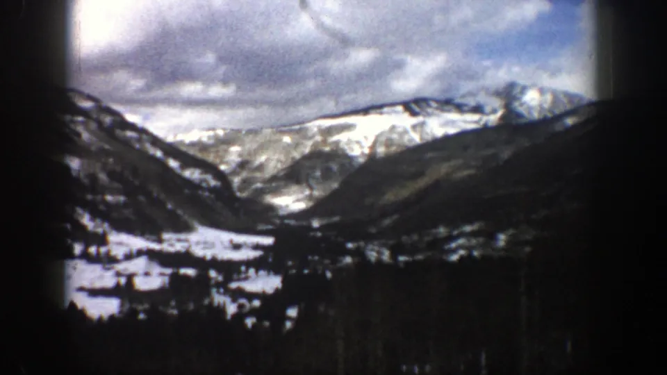 1961 Aspen-area home-movie frame showing a broad snowy valley, dark foreground trees, and white mountain ridges under heavy clouds.