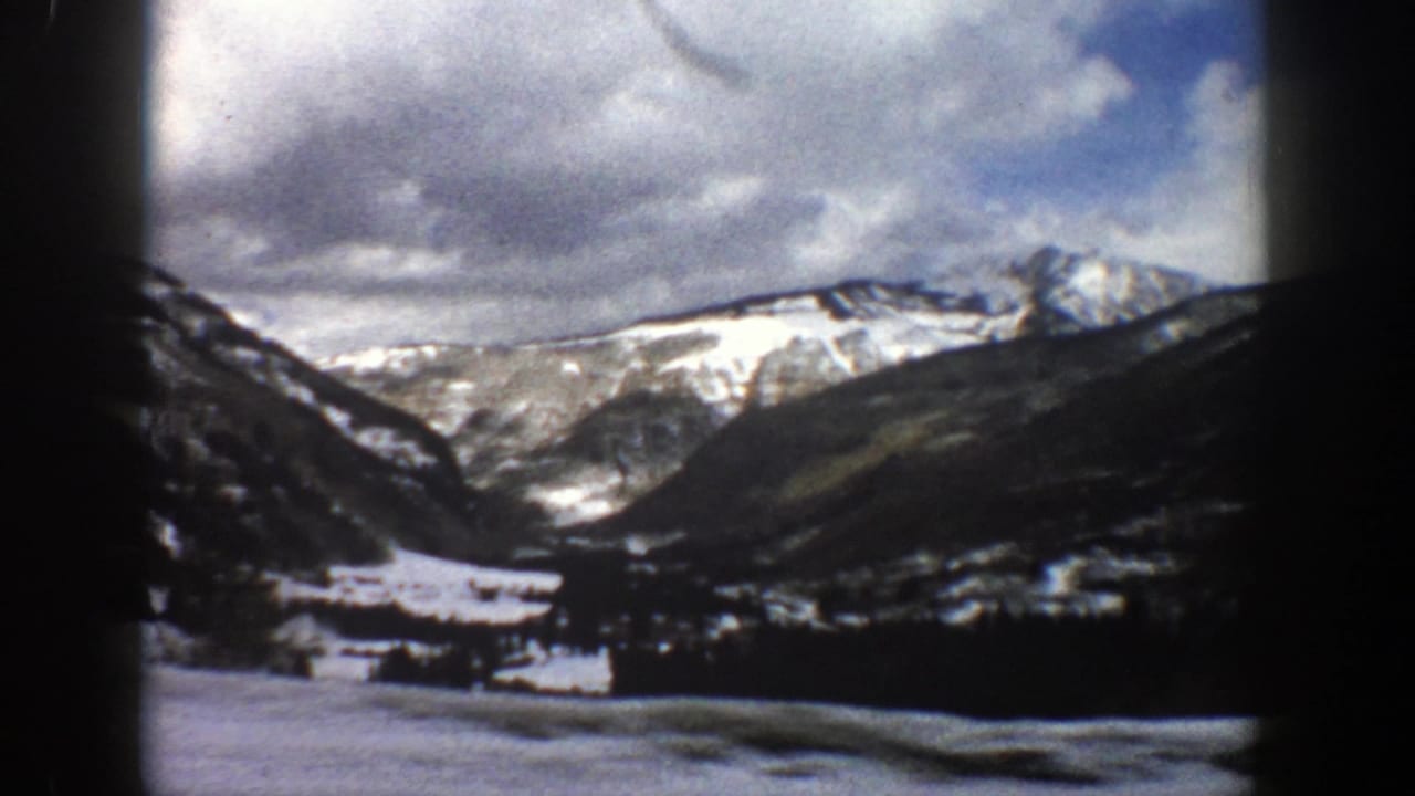 Snowy mountain valley beneath clouds, seen past dark roadside trees.