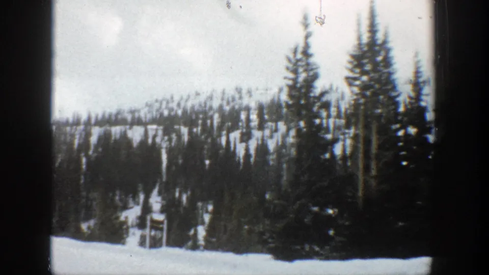 Aspen winter home-movie frame with foreground snowbank, dark evergreens, and a wooded snowy ridge beyond.
