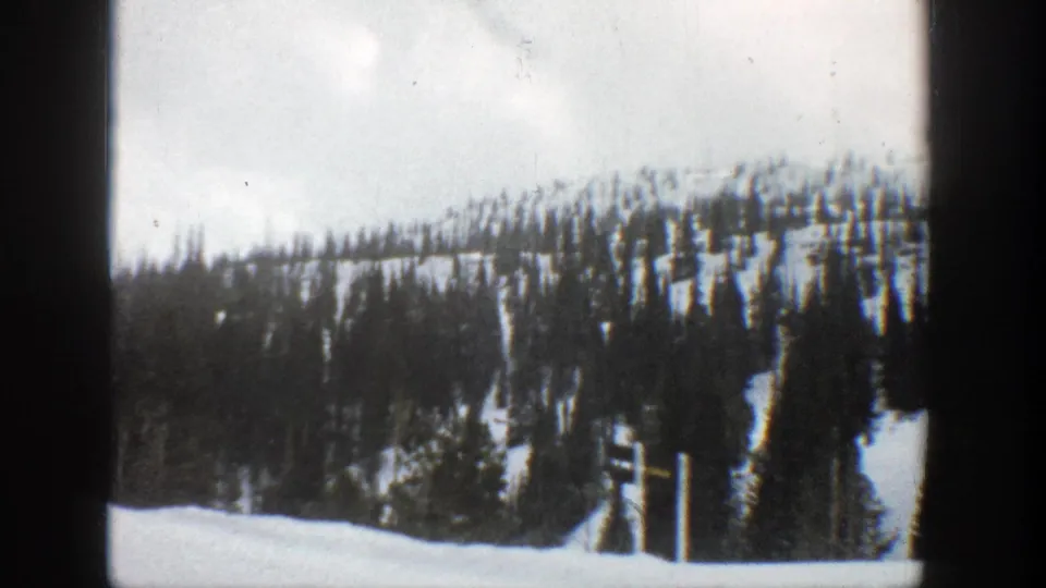 1961 Aspen 8mm frame of a snow-covered wooded slope, cloudy sky, and a small fence-like vertical at the lower right.