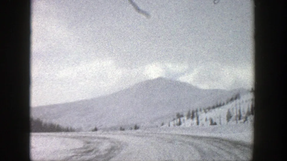 Forward vehicle-view frame of a snowy Aspen-area road, with tire tracks leading toward a hazy mountain ridge.