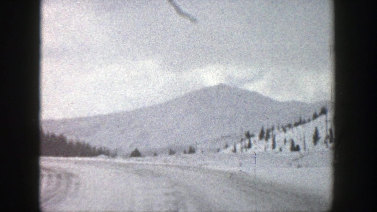 Snow-covered road curving through a winter field with distant mountains and scattered evergreens.