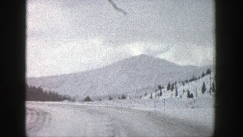 1961 Aspen, Colorado home-movie frame showing a snowy road, open field, tree-dotted hillside, and hazy mountain ridge under an overcast sky.