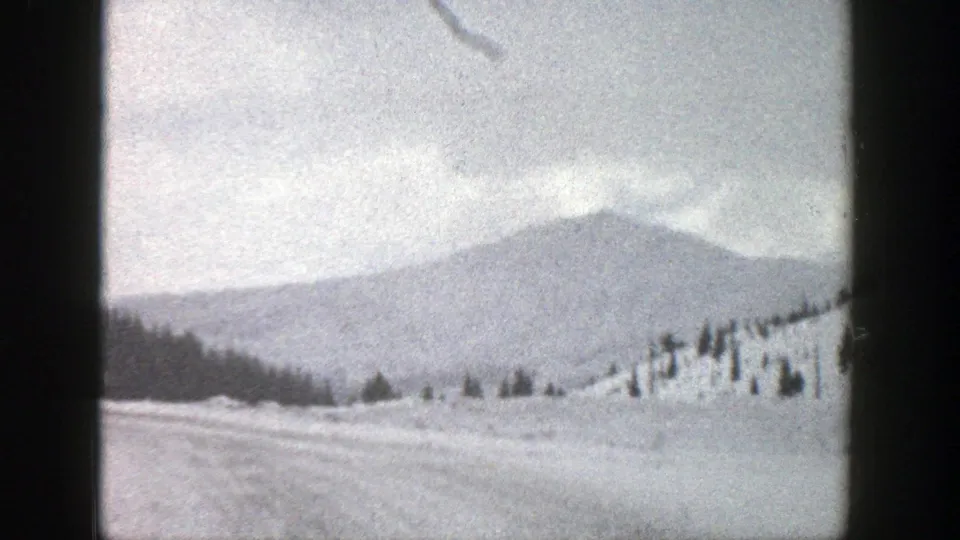 1961 Aspen, Colorado 8mm view of a snow-covered field and road edge with scattered evergreens and a soft distant mountain silhouette.