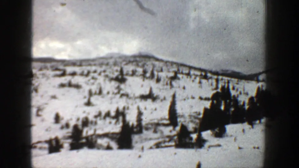 1961 Aspen, Colorado frame showing a wider snowy hillside, small evergreen clusters, low mountain ridge, and cloudy winter sky.