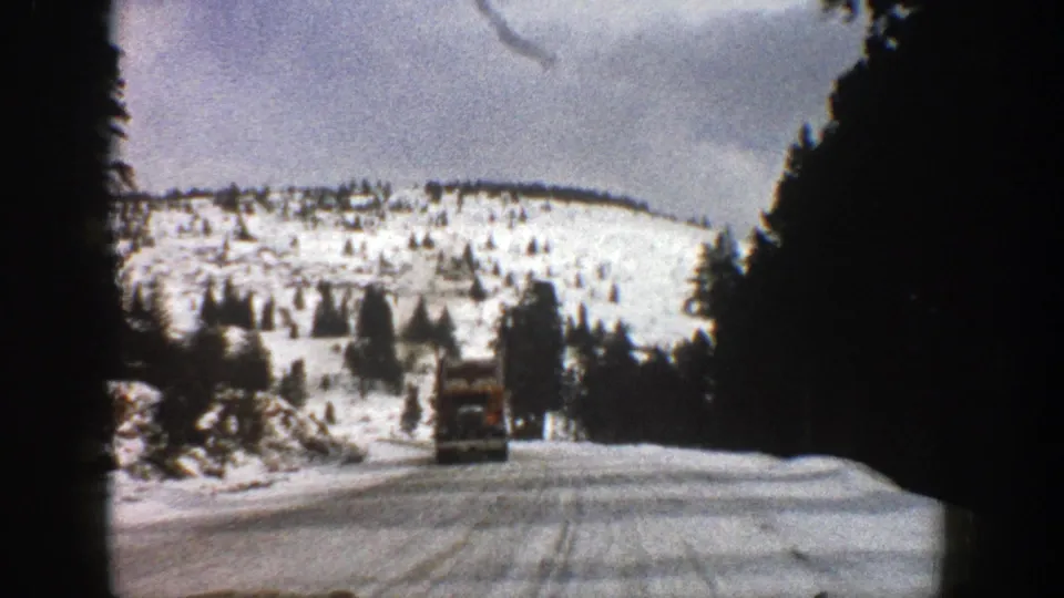 1961 Aspen-area winter road view with a red dump truck approaching between snowbanks and dark evergreen trees.