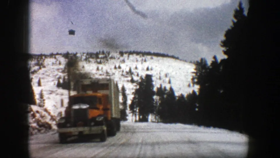 1961 Aspen-area 8mm frame showing a red dump truck passing close on a winter mountain road with snow and evergreens around it.