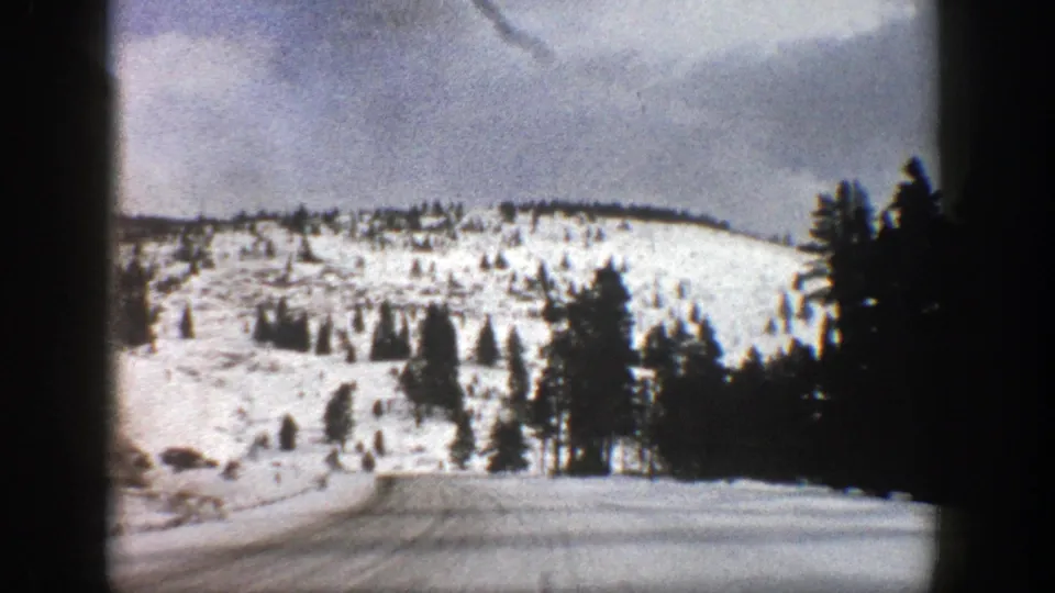 1961 Aspen-area winter road scene after the truck has passed, with a snowy roadway, evergreen stands, and a pale mountain slope ahead.