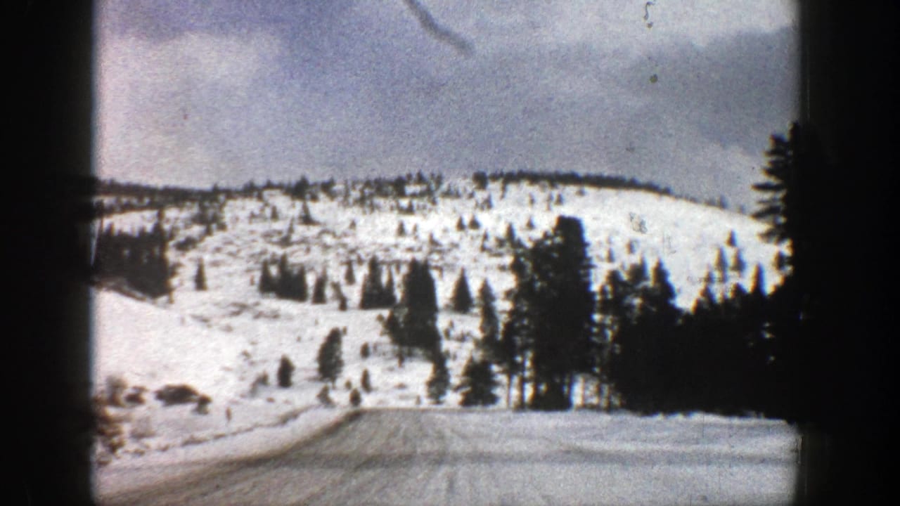 1961 Aspen, Colorado 8mm frame showing a snow-covered road approaching evergreen trees and pale mountain slopes.