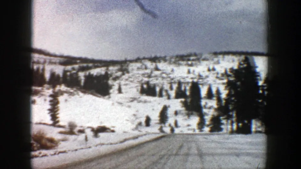1961 Aspen, Colorado home-movie frame with a snow-dusted road, evergreen clusters, and white hills ahead.