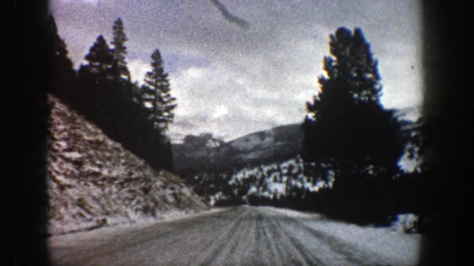 1961 Aspen, Colorado home-movie frame looking down a snowy road between dark evergreens and a steep roadside bank.