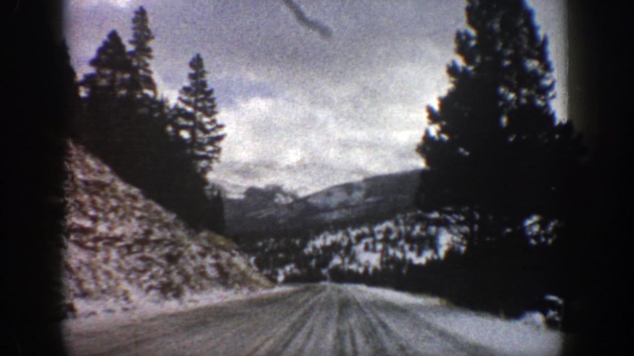 Snowy mountain road seen from a vehicle, with evergreen trees and distant cloudy peaks.