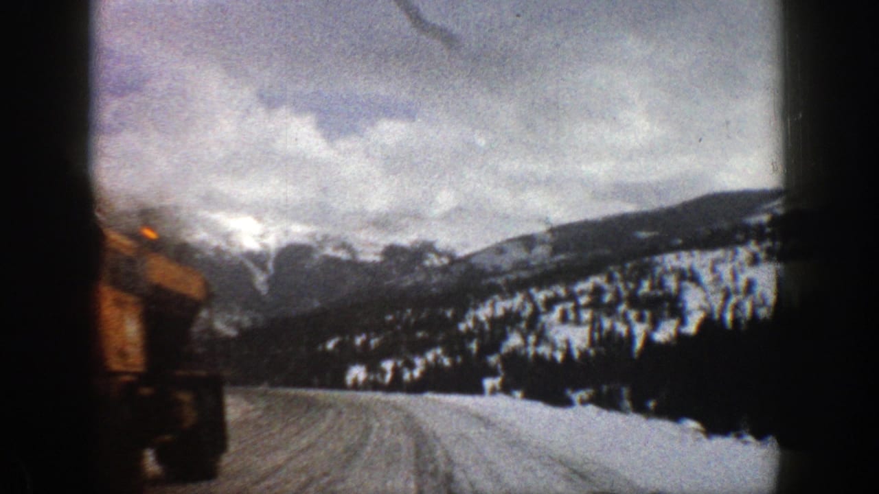 Snowy road beside mountain slopes with a road vehicle partly visible at left.