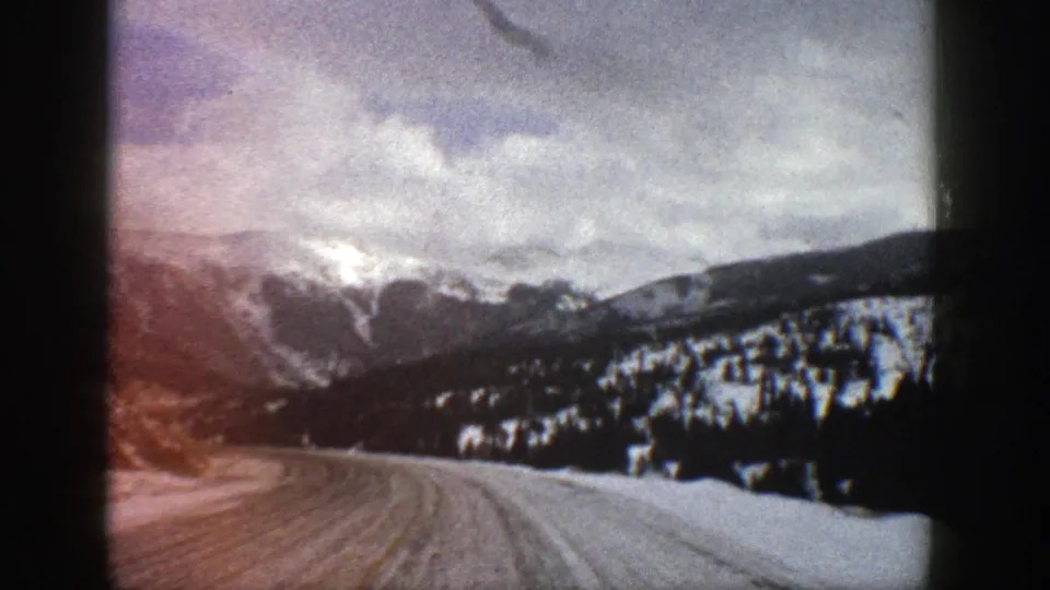 1961 Aspen-area 8mm view down a snow-cleared road toward evergreen slopes and clouded mountain ridges.
