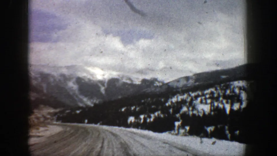 1961 Aspen-area 8mm frame with a snowy road foreground, evergreen-covered hillside, and soft cloudy mountain range beyond.