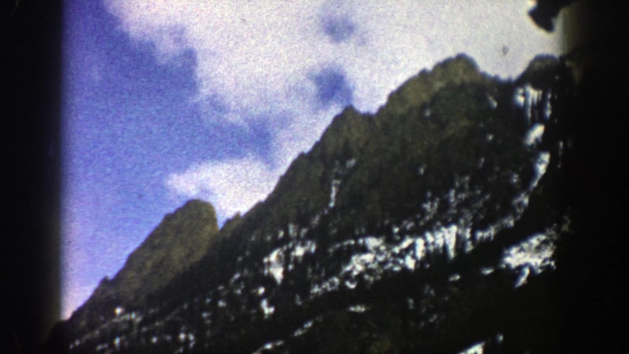 1961 Aspen-area 8mm frame showing rugged snow-dusted peaks, dark forested slopes, and bright clouds against blue sky.