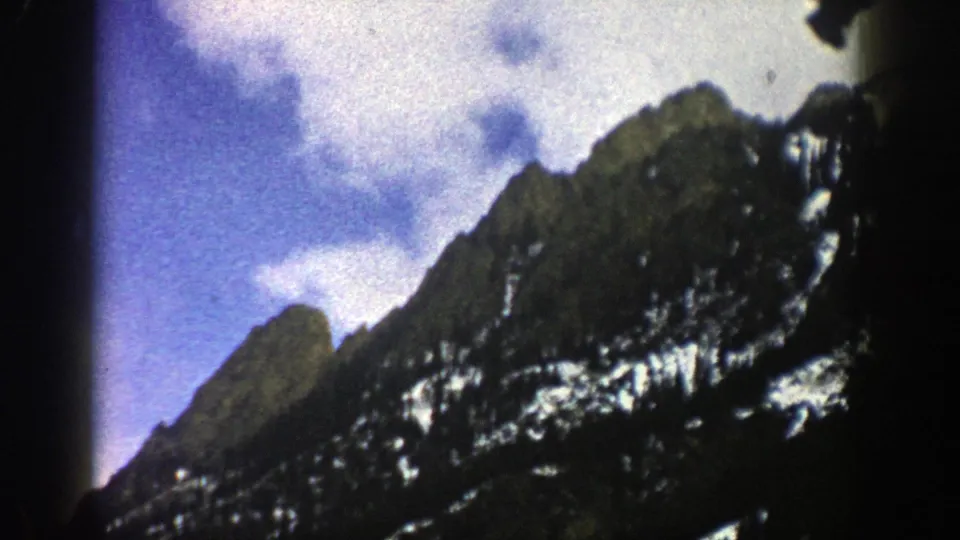 1961 Aspen-area 8mm frame showing rugged snow-dusted peaks, dark forested slopes, and bright clouds against blue sky.
