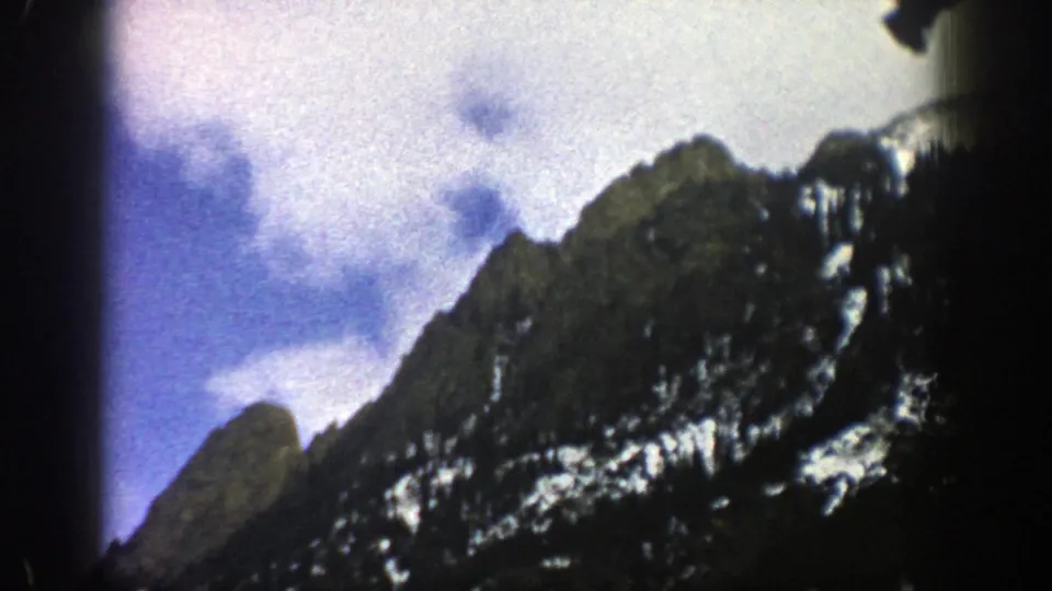 1961 Aspen-area mountain view with a jagged dark ridge, scattered snow patches, forested lower slope, and white clouds.
