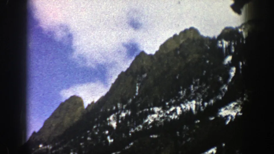 1961 Aspen-area 8mm frame of a dark forested mountain slope, streaks of snow, and bright clouds moving across blue sky.